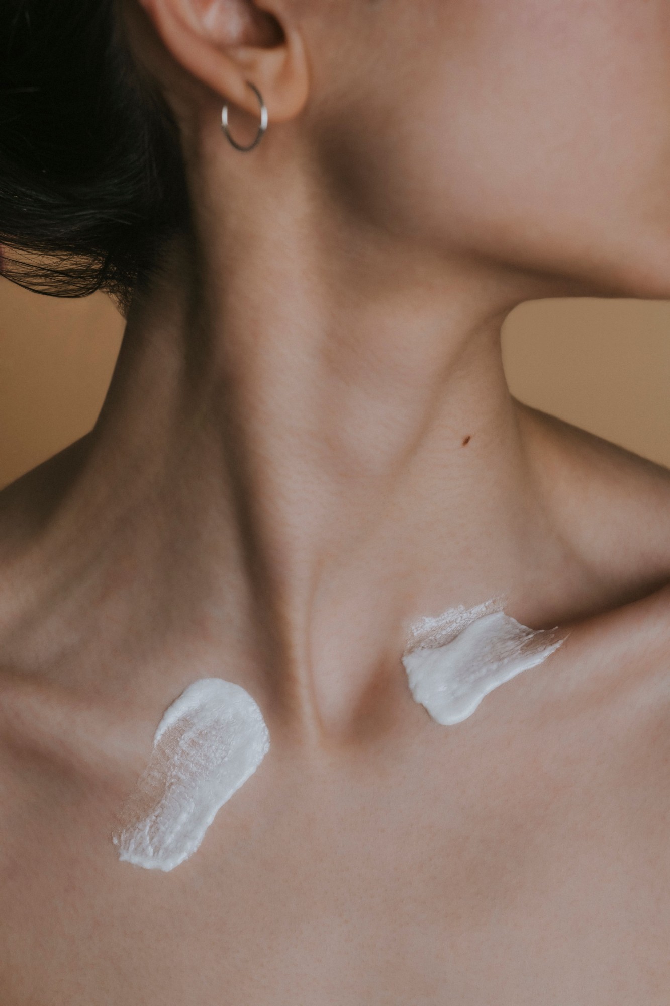 Two soft strokes of moisturizer on a person's collarbone, photographed against a warm neutral background.