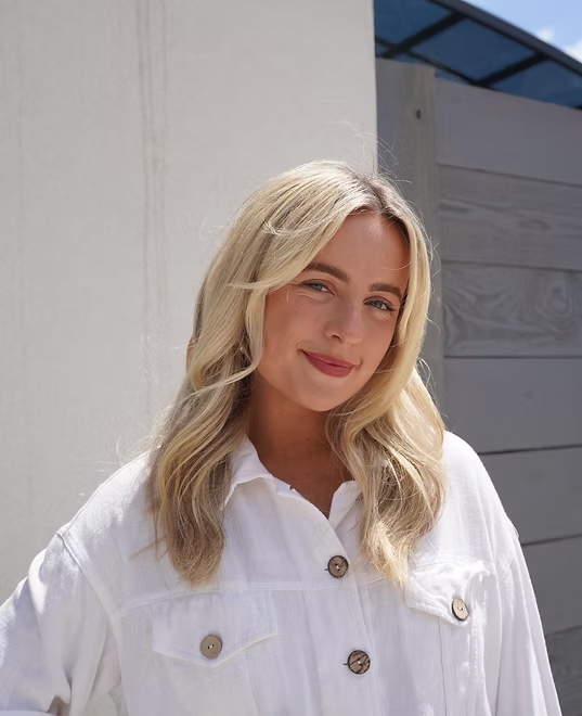 Olivia Jean, smiling in a white linen shirt in front of a sunlit white wall.